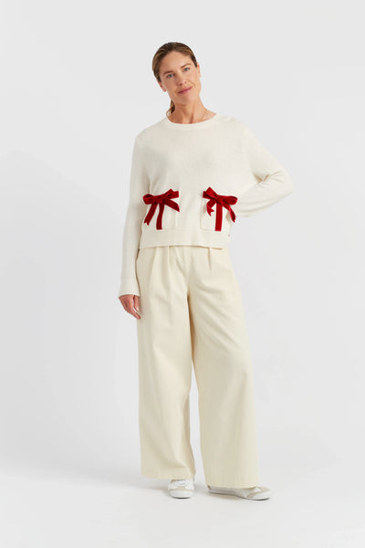 Woman wearing a cream sweater with red bow details and cream pants on a white background