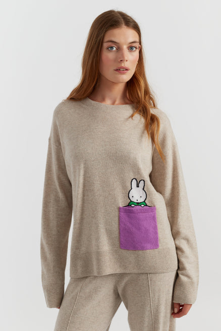Woman wearing a beige sweater with a cartoon character in a purple pocket on a white background