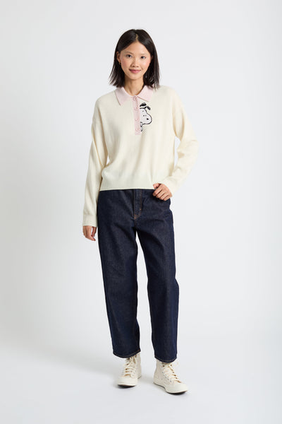 Person wearing a cream sweater with a logo and dark jeans on a white background