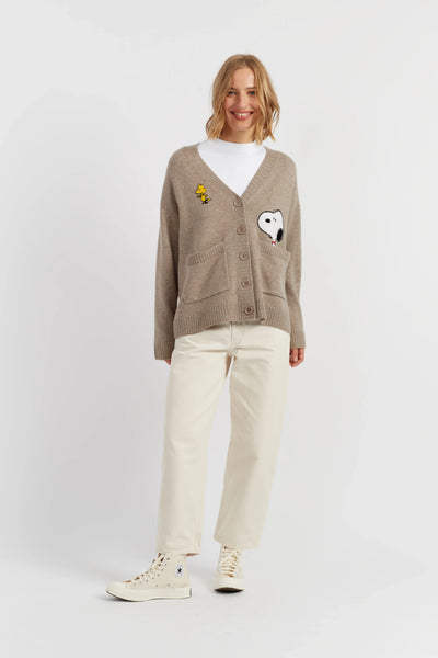 Woman wearing a brown cardigan with unique designs, white pants, and white sneakers on a light gray background